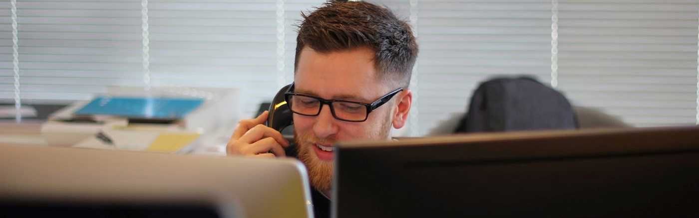 Mand med skæg, der snakker i telefon. Computerskærme dækker lidt af billedet.