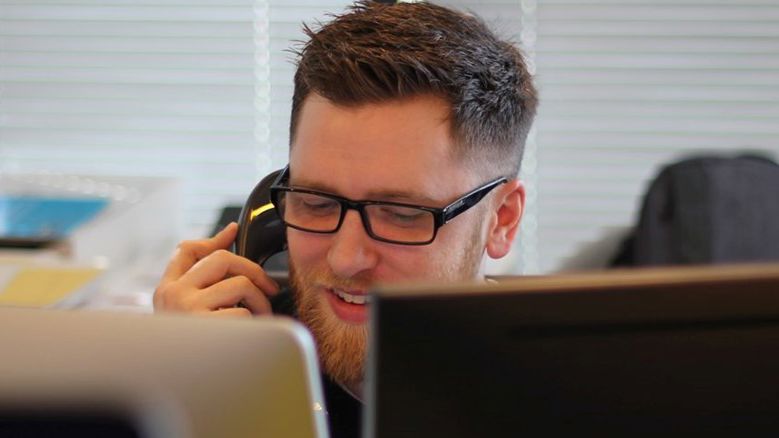 Mand med skæg, der snakker i telefon. Computerskærme dækker lidt af billedet.